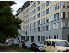Mehrstöckiges Bürogebäude mit klar gegliederten Fensterreihen und einer Fassade aus beige- und graufarbenem Stein. Im Vordergrund eine Straße mit parkenden Autos und ein Gehweg, der von Bäumen gesäumt ist.
