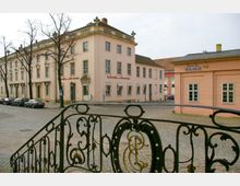 Historische Gebäude mit hellen Fassaden am gepflasterten Platz, darunter eine Gaststätte mit der Aufschrift "Gaststätte zur Ratswaage" und ein kleineres Gebäude mit der Aufschrift "Restaurant Waage". Im Vordergrund ein dekoratives, dunkles Metallgeländer.