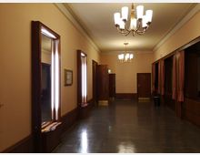 Ein eleganter Flur mit hellbeigen Wänden, glänzendem dunklen Holzboden und mehreren Hängeleuchten mit Milchglaslampen. An den Seiten befinden sich Holzrahmenfenster mit Spiegeln sowie Türen und Vorhänge aus dunklem Holz und Stoff in warmen Farbtönen.