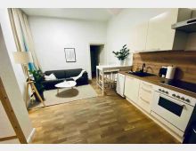 Offener Wohnbereich mit integrierter Küchenzeile in Weiß, Holzarbeitsplatte und dunkler Spüle sowie Herd und Backofen. Daneben stehen ein kleiner Esstisch mit Hockern und eine Sitzecke mit Sofa, rundem Couchtisch, Stehlampe und großem Fenster mit Vorhängen auf Holzboden.