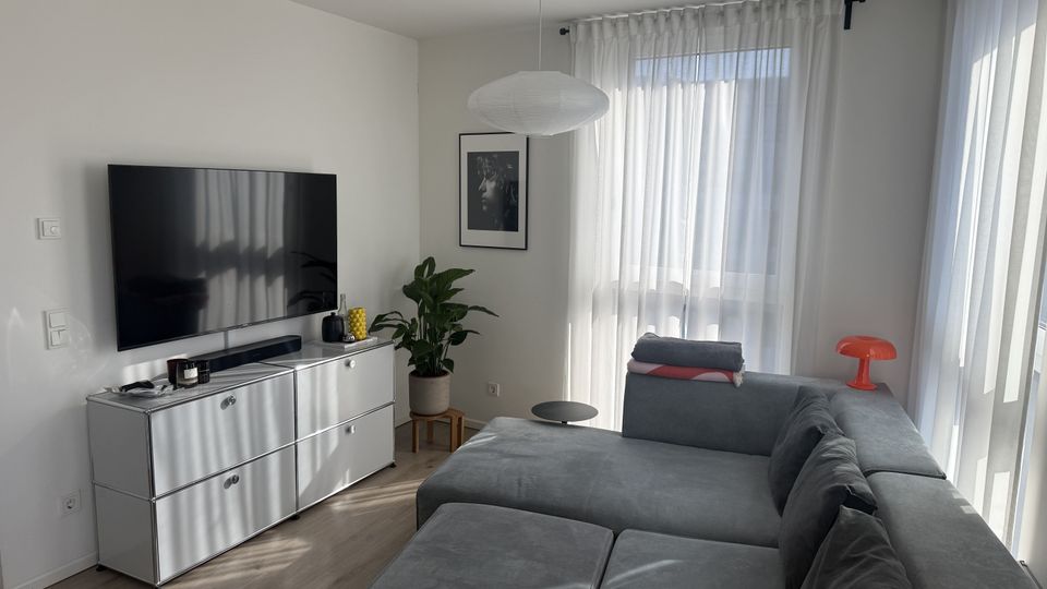 Helles, modern eingerichtetes Wohnzimmer mit großem grauen Ecksofa, wandmontiertem Flachbildfernseher über einem weißen Sideboard und einem runden Hängelampenschirm. Große Fenster mit transparenten Vorhängen lassen viel Tageslicht herein; eine Zimmerpflanze, ein gerahmtes Bild und eine kleine Tischlampe setzen Akzente.