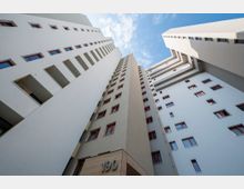 Blick von unten auf eine moderne, mehrstöckige Wohnanlage mit hellen Fassaden und roten Fensterrahmen, die symmetrisch angeordnet sind. Der Himmel ist blau mit wenigen Wolken, und der Eingangsbereich zeigt die Hausnummer 190.