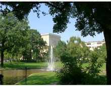 Ein grüner Park mit einem Teich, in dem ein Wasserbrunnen sprudelt, umgeben von Bäumen und Rasenflächen. Im Hintergrund sind mehrere mehrstöckige Gebäude zu sehen.