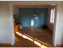Ein leerer Raum mit Holzboden und einer blauen Wand, beleuchtet durch Sonnenlicht. An der linken Wand steht ein Sideboard mit Pflanzen und Dekoration, rechts sind Fenster mit weißem Rahmen.
