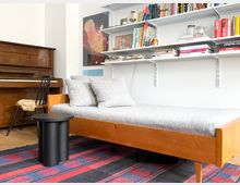 Helles Wohnzimmer mit Holz-Recamiere und grauen Kissen auf gemustertem Teppich, daneben ein schwarzer Beistelltisch. An der Wand befinden sich offene Regale mit Büchern und Gläsern; links steht ein Klavier neben einem Stuhl.