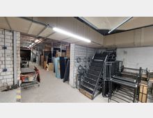 Lagerraum mit Betonboden und weiß gestrichenen Wänden, beleuchtet von langen Deckenleuchten. Der Raum enthält gestapelte Metallregale, Holzplatten, Metallrahmen und Kabelrollen, die entlang der Wände und auf dem Boden gelagert sind.