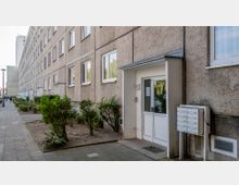 Mehrstöckiges Wohngebäude mit schlichter Putzfassade, rechteckigen weißen Fenstern und einem Hauseingang mit Glastür, daneben ein Briefkastenblock. Vor dem Gebäude verlaufen ein schmaler Grünstreifen mit Büschen und ein gepflasterter Gehweg.