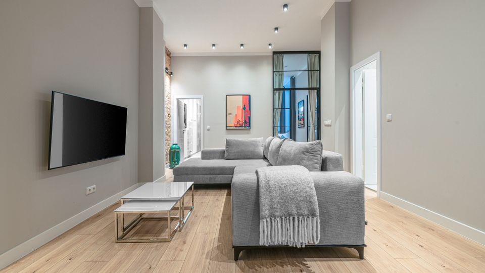 Modernes Wohnzimmer mit hellem Holzboden, grauen Wänden und hoher Decke mit Einbauspots, eingerichtet mit großem grauen Ecksofa und einem niedrigen Couchtisch. An der Wand hängt ein Flachbildfernseher; im Hintergrund führen Türen und eine verglaste Trennwand zu weiteren Räumen, ergänzt durch gerahmte Kunstwerke.