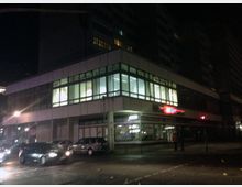 Eckgebäude in einer städtischen Umgebung bei Nacht, mit großen Glasfenstern im ersten Stock und einem beleuchteten roten Firmenschild im Erdgeschoss. Mehrere geparkte und fahrende Autos befinden sich auf der Straße davor.