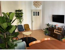 Wohnzimmer mit Holzboden, einer weißen Tür und einer an der Wand montierten schwarzen TV-Anlage. Im Vordergrund steht ein runder Holztisch, daneben befinden sich Zimmerpflanzen, zwei Sessel in Blau und Gelb, sowie ein kleines Couchtisch. Rechts steht ein Sideboard mit dekorativen Objekten und einem Retro-Radio. An der Decke hängt eine große, kugelförmige Pendelleuchte.