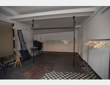 Fotostudio mit einfacher Einrichtung, bestehend aus einem grauen Hintergrund auf einem Ständer, Beleuchtungsausrüstung, einem Hocker, einem Kleidungsständer mit Kleiderbügeln und weißen Wandfliesen an der Rückwand. Der Boden ist dunkel mit einem schwarz-weiß karierten Bereich.