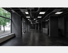 Großzügiger Innenraum mit dunklem, minimalistischem Design, einer offenen Decke mit sichtbaren Leitungen und rechteckigen Leuchtstofflampen. Auf der linken Seite sind große Fenster und eine Glastür, die den Blick auf Bäume draußen ermöglicht.