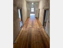 Ein schmaler Flur mit frisch verlegtem Holzboden, grob verputzten Wänden und einer einfachen Glühbirne an der Decke. Am Ende des Flurs befindet sich ein Fenster, das Tageslicht hereinlässt, und es gehen Türen zu angrenzenden Räumen ab.