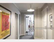 Ein heller Flur mit beigen Wänden, an denen Bilder und Kunstwerke, darunter ein Keith-Haring-Poster, hängen. Am Ende des Flurs befindet sich ein Raum mit Staffeleien und Malutensilien, der durch eine helle Fensterfront beleuchtet wird.