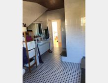 Ein Badezimmer mit hellen Wandfliesen und einem Boden aus schwarz-weißem Mosaikmuster. Im Vordergrund stehen ein Holzregal und ein Doppelwaschbecken mit Spiegel, im Hintergrund führt eine geöffnete Tür zu einem angrenzenden Raum mit Holzboden.