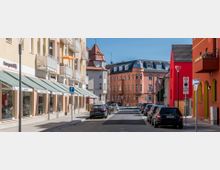 Straßenszene mit mehrstöckigen Wohn- und Geschäftshäusern in kräftigen Farben, links mit Balkonen und Markisen, rechts mit roten Fassaden. Im Hintergrund steht ein orangefarbenes Eckgebäude mit Mansardendach, parkende Autos säumen die Straße.