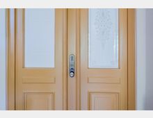 Zweiflügelige Holztür in hellbeigem Anstrich mit profilierten Kassetten und zwei hohen Milchglasfeldern mit ornamentaler Ätzung. In der Mitte befindet sich ein Metallbeschlag mit Zahlencode-Tastatur und runder Griffgarnitur.