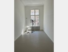 Schmaler, weiß gestrichener Raum mit einem großen, bogenförmigen Fenster, das auf ein gegenüberliegendes Gebäude blickt. Im Raum stehen eine Stehlampe, ein kleiner Beistelltisch, eine weiße Tischlampe und ein schwarzer Kleiderständer; darunter ein grauer Fußboden und ein Heizkörper unter dem Fenster.