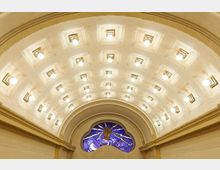 Gewölbedecke eines historischen Innenraums mit zahlreichen eingelassenen Lichtern in symmetrischer Anordnung. Unterhalb der Decke befindet sich ein buntes Glasfenster mit einer Darstellung einer erhobenen Hand und strahlenförmigen Mustern.