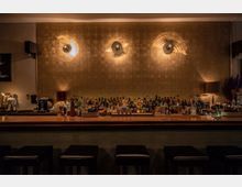 Eine stilvoll eingerichtete Bar mit einer langen Theke aus Holz, dahinter eine Vielzahl von Spirituosenflaschen und Barutensilien. Die Wand im Hintergrund besteht aus goldfarbenen Kacheln, die von warmem Licht beleuchtet werden; davor befinden sich schwarze Barhocker.