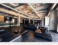 Industrieller Innenraum mit Sichtbetondecke, freiliegenden Lüftungsrohren und schwarzen Ledersofas. Im Hintergrund befindet sich eine lange Theke, ergänzt durch Vorhänge und schlichte Dekoration.