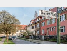 Wohnstraße mit zweigeschossigen Reihenhäusern in Pastelltönen, roten Dächern und Fensterläden; links ein Baum und parkende Autos, rechts ein Straßenschild mit der Aufschrift „Thanner Pfad“.