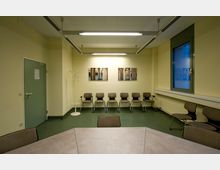 Wartezimmer mit gelb gestrichenen Wänden, mehreren gepolsterten Stühlen in einer Reihe entlang der hinteren Wand, einem weißen Kleiderständer und zwei gerahmten Wandbildern. Links befindet sich eine grüne Tür, rechts ein großes Fenster mit dunkelgrünem Rahmen, vorne ein Tischabschnitt.