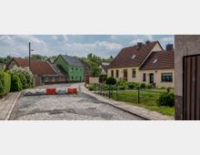 Eine gepflasterte, kurvige Straße in einem Dorf mit kleinen Einfamilienhäusern und roten sowie grünen Fassaden. Im Vordergrund sind Absperrungen auf der Straße, daneben Gehwege und gepflegte Gärten mit Rasen und Büschen.