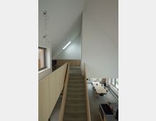 Innenansicht eines modernen Hauses mit offenen Wohnbereichen. Im Vordergrund führt eine gerade Betontreppe mit Holzgeländern in ein Obergeschoss, während im Hintergrund ein Essbereich mit einem langen Holztisch und mehreren Stühlen sowie eine offene Küche zu sehen sind.