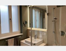 Modernes Badezimmer mit einer Dusche mit Glastür, einem weißen Waschbecken auf einem Schrank, beleuchtetem Wandspiegel und einem Fenster mit Milchglas. Handtücher hängen an einem Handtuchhalter unter dem Fenster.