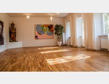 Ein heller Raum mit Holzparkettboden, weißen Wänden und großen Fenstern, durch die Tageslicht einfällt. An der Wand hängen ein farbiges abstraktes Gemälde und eine dekorative Statue; rechts steht eine große Zimmerpflanze in einem weißen Topf.