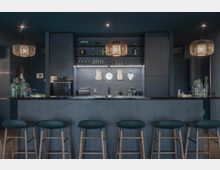 Moderne Küche mit einer dunklen Arbeitsfläche und integrierter Bar mit fünf Barhockern. Über der Arbeitsfläche hängen dekorative Hängelampen, und im Hintergrund befinden sich Einbauschränke, offene Regale mit Geschirr und Wanddeko.