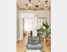 Wohnzimmer mit modernem, skandinavischem Design, bestehend aus einem grünen Ledersessel, Holzboden im Fischgrätmuster und einer Hängelampe mit mehreren Leuchtarmen. Eine offene weiße Doppeltür führt in einen angrenzenden Raum, während rechts große Zimmerpflanzen neben einem Glaselement stehen.