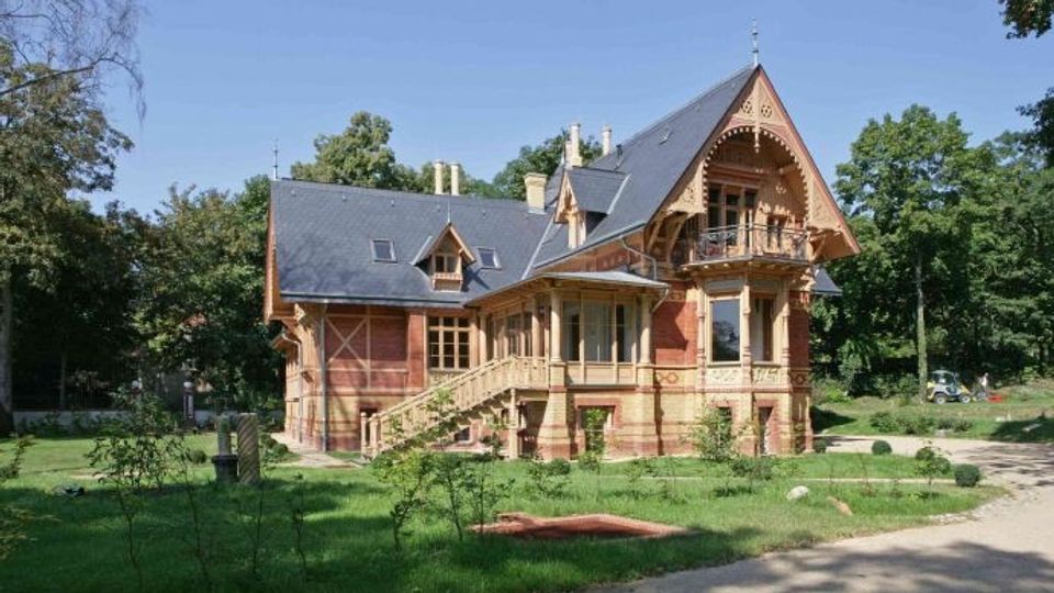 Historische Villa im Park, mit steilem Schieferdach, Ziergiebeln und reich verzierten Holz- und Backsteinfassaden samt Balkon und Veranda. Das Gebäude steht auf einer Rasenfläche mit Wegen und umgebenden Bäumen.