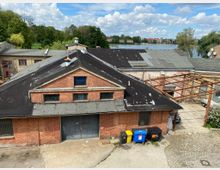 Ein Industriegebäude aus rotem Backstein mit schrägem, schwarzem Dach, großen Metalltoren und kleinen Fenstern. Im Vordergrund stehen farbige Mülltonnen, im Hintergrund ist eine grüne Landschaft mit Bäumen und ein See sichtbar.