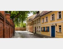 Pflastersteinstraße in einer historischen Altstadt mit bunten, zweistöckigen Häusern; links eine Backsteinmauer mit Straßenlaternen, und im Hintergrund große grüne Bäume.