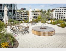 Eine modern gestaltete Dachterrasse mit hellen Steinplatten, zahlreichen Pflanzenbeeten, mehreren Holzbänken und Metalltischen mit zugehörigen Stühlen. Im Hintergrund sind umliegende Wohn- und Bürogebäude zu sehen, zusammen mit geschlossenen Sonnenschirmen.