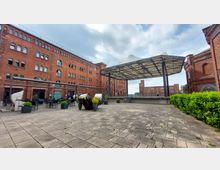 Großer, gepflasterter Innenhof eines ehemaligen Backstein-Speichergebäudes, umgeben von roten Ziegelbauten mit Rundbogenfenstern. In der Mitte steht eine moderne, überdachte Bühne, rechts im Bild sind Büsche und der Eingang zu einer Kirche mit der Aufschrift „Die 95 Thesen von Wittenberg“ zu sehen.