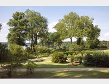 Weitläufiger Park mit großen, alten Bäumen, gepflegten Blumenbeeten und Rasenflächen; dahinter ist ein Fluss oder See sichtbar, umgeben von dichtem Grün. Der Himmel ist klar mit wenigen Wolken, und Spazierwege durchziehen die Landschaft.