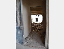 Blick durch eine Türöffnung in einem verlassenen, zerfallenen Gebäude mit Schutthaufen auf dem Boden und teilweise eingestürzten Wänden. Der Raum dahinter zeigt beschädigte Strukturen und Lichteinfall durch ein geöffnetes Fenster.