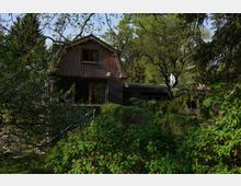 Holzhaus mit Giebeldach und großen Fenstern, umgeben von dichter Vegetation und Bäumen in einem Garten. Auf dem Flachdach eines angrenzenden Gebäudeteils steht eine weiße Skulptur, und der Hintergrund zeigt weitere Bäume.