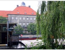 Historisches Gebäude mit heller Fassade und rotem Ziegeldach, im Hintergrund eines Hafengebiets. Im Vordergrund befinden sich Zweige einer Weide und ein Teil eines Schiffes mit der Aufschrift „H. Schulz Hamburg 865 L67 B8.2“.