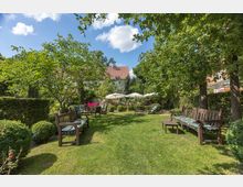 Ein gepflegter Garten mit einer Rasenfläche, umgeben von Bäumen und Büschen. Mehrere Holzbänke und Liegen mit grünen Auflagen stehen verteilt im Garten, während im Hintergrund weiße Sonnenschirme Schatten spenden; dahinter ist ein Gebäude mit rotem Ziegeldach sichtbar.