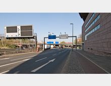 Straßenansicht mit mehreren Fahrspuren, Verkehrsschildern und einer Brücke im Hintergrund. Rechts befindet sich ein großes Gebäude mit pinkfarbener Steinfassade und Fenstern, links führen Leitplanken entlang der Straße; im Hintergrund sind Bäume und weitere Verkehrswege sichtbar.