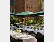 Hof eines Backsteingebäudes mit gedeckten Tischen für eine Veranstaltung im Freien. Die Tische sind mit weißen Decken, Blumenarrangements und elegantem Gedeck dekoriert, umgeben von grünen Sonnenschirmen und Pflanzen.