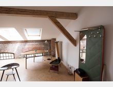 Dachwohnung mit sichtbaren Holzbalken, einer Backsteinwand und großen Dachfenstern, die Tageslicht einlassen. Der Raum enthält einen Schreibtisch, eine Stehlampe, einen kleinen Tisch und eine Wandgarderobe mit Spiegel.