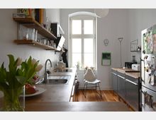 Moderne Küche mit Holzarbeitsplatten, offener Regalbödenwand und großem Bogenfenster, das Tageslicht in den Raum bringt. Im Vordergrund sind eine Vase mit weißen Tulpen und ein Obstteller zu sehen, während helle Wände und schwarze Küchenschränke den Raum definieren.