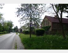 Straßenszene in einer ländlichen Gegend mit einem gepflasterten Bürgersteig, grünen Rasenflächen und Bäumen. Auf der rechten Seite steht ein Backsteingebäude mit roten Dachziegeln und bewachsenen Fassaden, während links die Straße von Hecken flankiert wird.