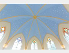 Blick nach oben auf das blaue, sternenverzierte Gewölbe einer gotischen Kirche mit spitzbogigen Fenstern, die bunte Glasornamente zeigen. Die Architektur wird von goldenen Rippen betont.