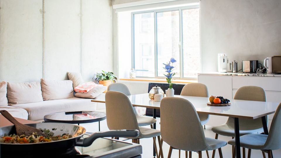 Heller, moderner Wohn- und Essbereich mit großem Fenster, hellem Sofa und einem rechteckigen Esstisch mit mehreren gepolsterten Stühlen. Rechts befindet sich eine schlichte weiße Küchenzeile mit kleinen Küchengeräten und Arbeitsfläche.